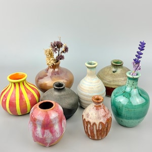 May include: A collection of eight small ceramic vases in various shapes and finishes. The set includes pieces with bright yellow stripes, pink and white drip glazes, a matte charcoal finish, a light blue ribbed texture, and a vibrant green glaze.