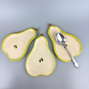 May include: Three pear-shaped ceramic dishes with a speckled cream interior and a bright lime green rim. Each dish features a small brown stem at the top and two dark brown seed shapes in the center. A silver metal spoon rests on one dish.