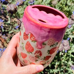 May include: A ceramic tumbler with a pink, dripped glaze and a strawberry pattern. The top rim is a darker pink, and the interior is a deep red. The tumbler is cylindrical and appears to be handmade.