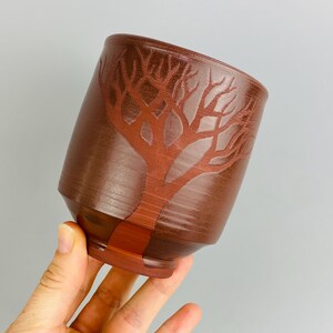May include: A brown ceramic cup with a tree design carved into the surface. The tree is a lighter shade of brown, creating a contrast against the darker background. The cup has a slightly angled shape.