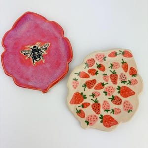 May include: Two irregular, hand-formed ceramic dishes. One dish features a vibrant, textured pink glaze with a central black and white bumblebee illustration. The second dish is cream-colored and decorated with a repeating pattern of red and pink strawberries.