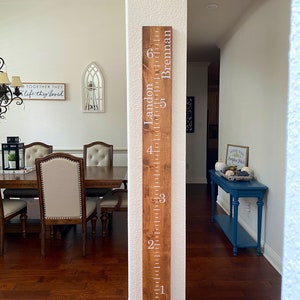 May include: A tall, rectangular wooden growth chart ruler mounted vertically on a wall. The board features a dark wood stain with white painted markings for feet from one to six, and the names Landon and Brennan printed in white text.