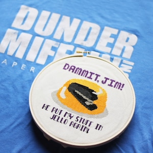 Op de afbeelding: Een borduurring met kruissteek met een ontwerp van gele Jell-O met een zwarte nietmachine en de tekst "DAMMIT, JIM! HE PUT MY STUFF IN JELLO AGAIN." De ring ligt op een blauw t-shirt met "Dunder Mifflin Paper".