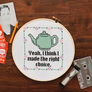 以下が含まれることがあります： 緑色のティーポットと「Yeah, I think I made the right choice.」という文字が書かれた白い刺繍枠。枠は木製の表面に置かれ、オレンジ色のホットソースのパケットが2つと人物の写真が置かれている。