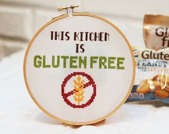 Può includere: Un cerchio da ricamo bianco con il testo "This kitchen is gluten free" e un cerchio rosso con una spiga di grano barrata. Sullo sfondo, una busta di cracker per sandwich al burro di arachidi senza glutine.