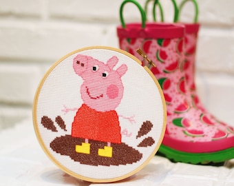 May include: A cross-stitch hoop featuring a pink pig in a red dress splashing in a mud puddle. The hoop is next to a pair of pink rain boots with a watermelon pattern and green soles.