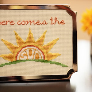 Könnte beinhalten: Ein gerahmtes Kreuzstichbild mit einer gelben Sonne, die über einem grünen Hügel aufgeht. Der Text "here comes the" ist in Orange über der Sonne gestickt.