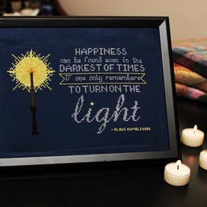 May include: Framed cross-stitch art with the quote "Happiness can be found even in the darkest of times...to turn on the light" by Albus Dumbledore. The artwork features a yellow wand and is set against a dark blue background. Three lit candles are in the foreground.