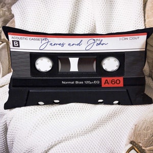 Puede incluir: Almohada de cinta de casete en blanco y negro con el texto "Acoustic Cassette" y "James and John" escrito en tinta azul. La almohada tiene una etiqueta roja y blanca con el texto "A 60" y "Normal Bias 120µs EQ".