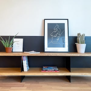 Könnte beinhalten: Ein Holzregal mit schwarzen Metallstützen. Das Regal hat zwei Ebenen und ist mit einem Schwarz-Weiß-Linienkunst-Druck, einer Topfpflanze und einer Topfaloe-Vera-Pflanze dekoriert. Das Regal ist auch mit Büchern und Zeitschriften dekoriert.