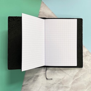 May include: A black leather-bound notebook with a gridded paper interior. The notebook is open and lying flat on a light grey surface.