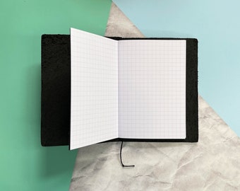 May include: A black leather-bound notebook with a gridded paper interior. The notebook is open and lying flat on a light gray surface.