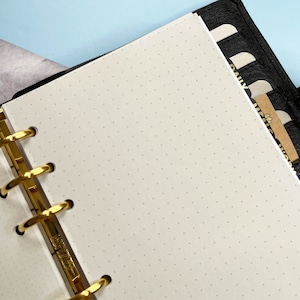 May include: A white, dotted, paper planner insert with gold rings. The insert is in a black leather planner with tabs labelled "Daily", "Weekly", and "Monthly".