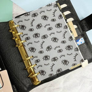 May include: A clear plastic planner insert with a black and white eye pattern. The insert is designed to fit a standard size planner and has tabs for different sections, including "Months", "Daily", "Meals", "Work", and "Important".