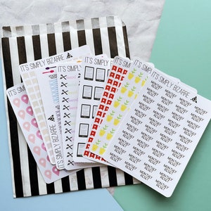 Op de afbeelding: Een set kleurrijke planner stickers met verschillende ontwerpen, waaronder een boek, een schaar, een plant en een locatiepin. De stickers zijn verpakt in een zwart-wit gestreepte papieren zak. De tekst "IT'S SIMPLY BIZARRE" is op de stickers gedrukt.