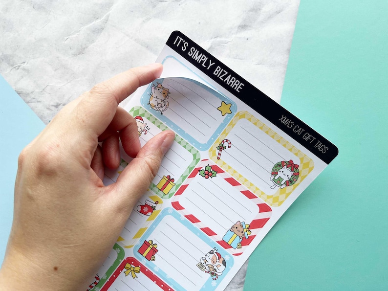 May include: Sheet of Christmas cat gift tags with various designs, including a cat in a wreath, a cat with a present, and a cat in a Santa hat. The tags have a white background with colorful borders and the text "It's Simply Bizarre" and "Xmas Cat Gift Tags".