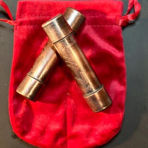 Puede incluir: Dos tubos de metal de color cobre con diseños grabados, descansando dentro de una bolsa de terciopelo rojo.