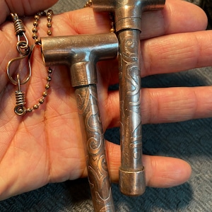 Può includere: Due ciondoli in tubo di rame con intricati disegni a spirale. I ciondoli sono attaccati a una catena argentata.