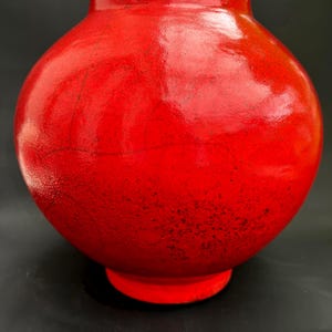 Könnte beinhalten: Eine leuchtend rote Keramikvase mit rundem Korpus und leicht ausgestelltem Rand. Die Vase hat eine glänzende Oberfläche und einen kleinen, kreisförmigen Sockel. Die rote Farbe ist satt und gleichmäßig, mit subtilen Farbnuancen, die dem Stück Tiefe verleihen. Diese dekorative Vase eignet sich für die Inneneinrichtung.