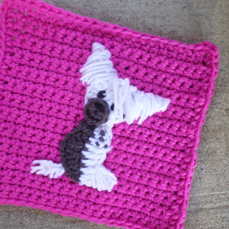 May include: A crocheted square with a white and brown dog applique. The square is pink and the dog is standing on its hind legs.