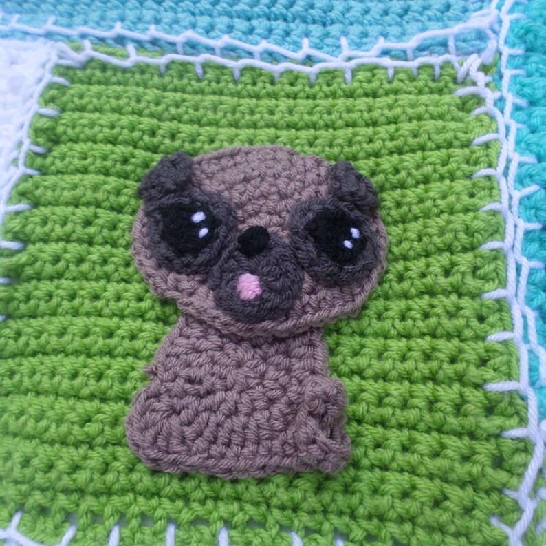 May include: A crocheted green square with a brown crocheted pug in the center. The pug has black eyes and a pink tongue.