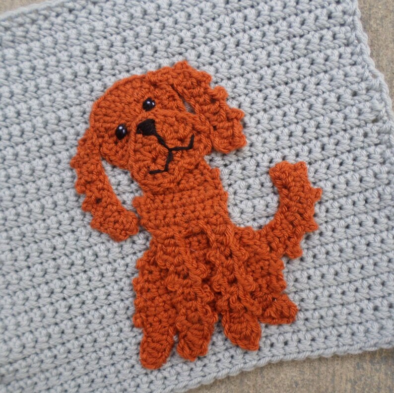 K&ouml;nnte beinhalten: Eine geh&auml;kelte orangefarbene Hundefigur auf einem grauen geh&auml;kelten Hintergrund. Der Hund hat schwarze Augen und eine schwarze Nase.