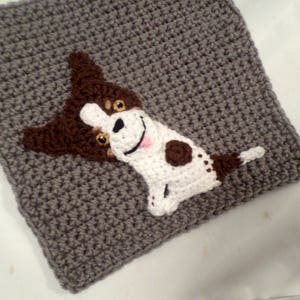 May include: A crocheted gray square with a brown and white dog applique. The dog has brown ears, brown spots, and a white face with black eyes and a pink tongue.