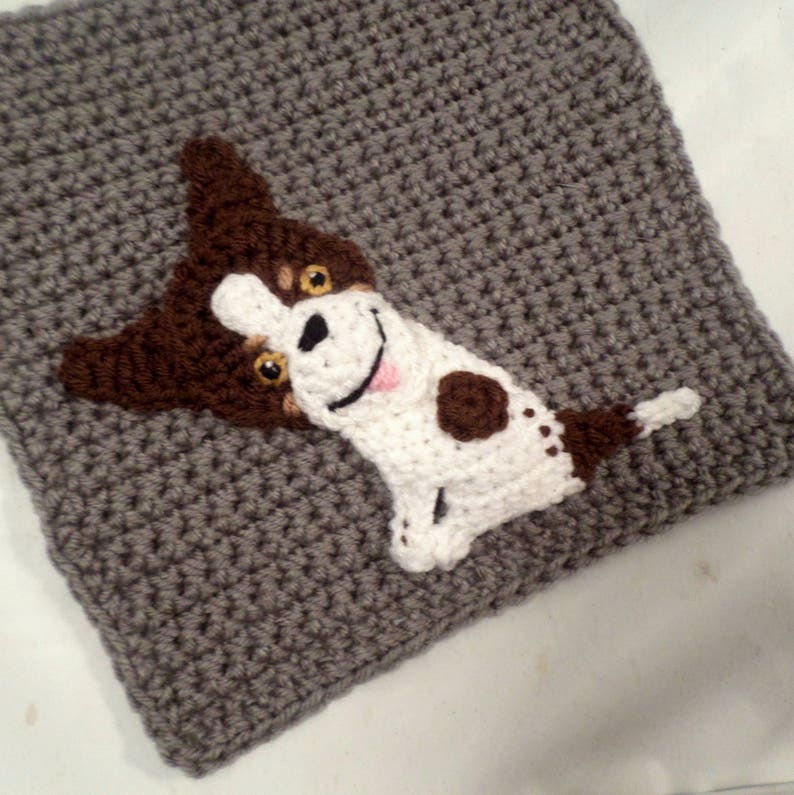 May include: A crocheted gray square with a brown and white dog applique. The dog has brown ears, brown spots, and a white face with black eyes and a pink tongue.