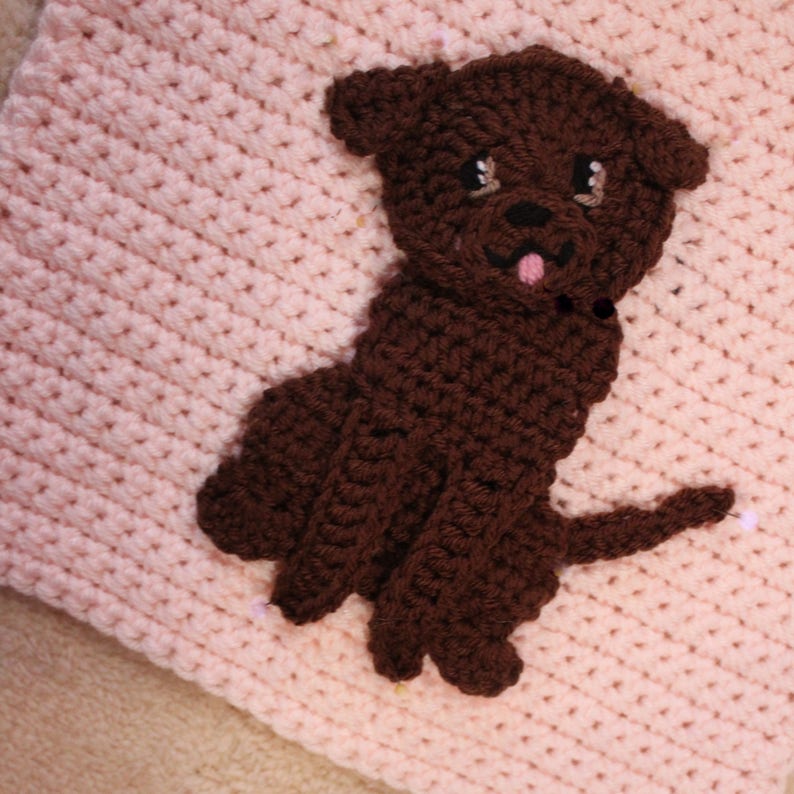 May include: A brown crocheted dog applique with a pink crocheted background. The dog has a white tongue sticking out and black eyes.