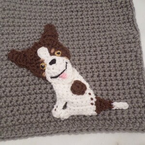 May include: A crocheted gray blanket with a brown and white dog applique. The dog is sitting with its tongue sticking out and has brown spots.