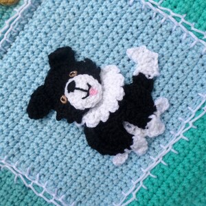 Puede incluir: Una manta de beb&eacute; de ganchillo con un perro de ganchillo blanco y negro en el centro. La manta est&aacute; hecha de hilo azul, verde y blanco.