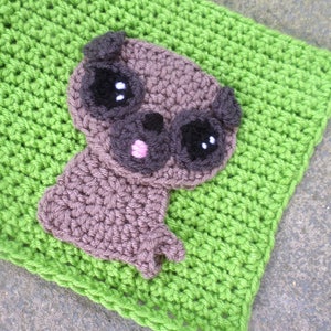 May include: A crocheted brown pug with black eyes and a pink nose, sitting on a green crocheted blanket.