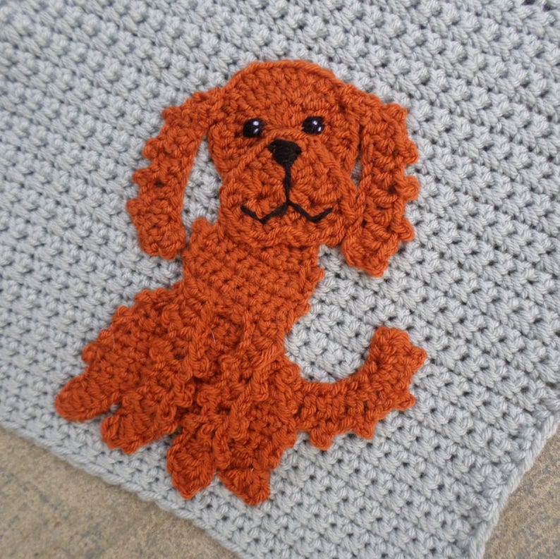 K&ouml;nnte beinhalten: Eine geh&auml;kelte Decke mit einer braunen geh&auml;kelten Hundefigur. Der Hund hat schwarze Augen und eine schwarze Nase.