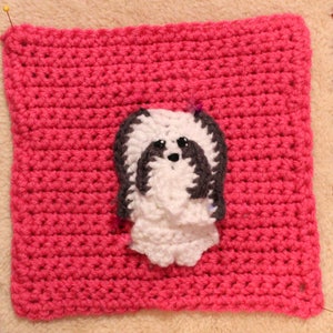 May include: A pink crocheted square with a white crocheted dog applique in the center. The dog has black ears and a black nose.