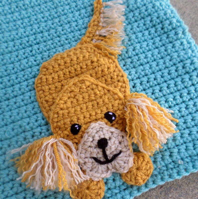 Puede incluir: Una aplicaci&oacute;n de perro de crochet en tonos de amarillo mostaza y crema, sobre un fondo azul claro. El perro tiene una expresi&oacute;n feliz y est&aacute; tumbado con la cola enroscada.