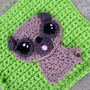 May include: A brown crocheted pug dog with black eyes and a pink nose, sitting on a green crocheted background.