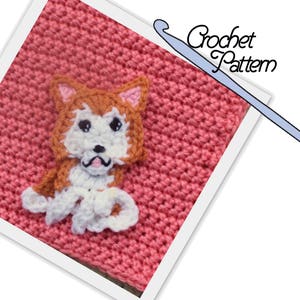 May include: A crochet pattern for a cute dog applique. The dog is brown and white with a pink nose and black eyes. The pattern is on a pink crocheted background.