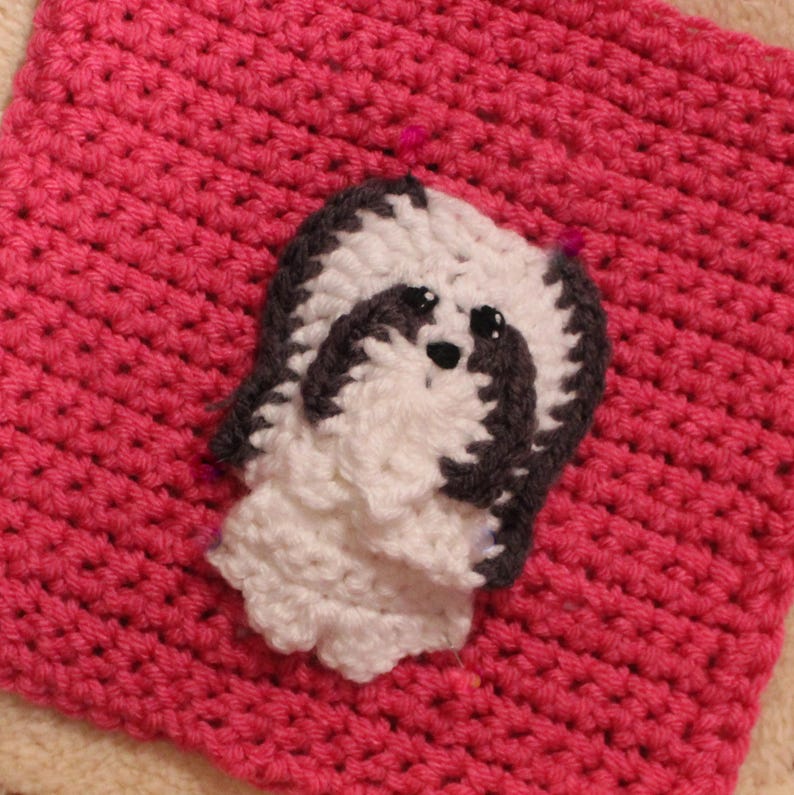 May include: A white crocheted dog with black accents is pinned to a pink crocheted square.