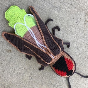 May include: A large, handmade crochet insect in shades of brown, tan, black, red, and green. The insect has large wings, a segmented body, and detailed features. The crochet work is visible, showcasing the texture and craftsmanship of the piece.