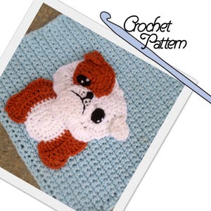 May include: A crochet pattern for a baby blanket with a cute brown, white, and orange bulldog applique. The blanket is a light blue color and the crochet hook is visible in the image.