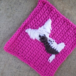 May include: A pink crocheted square with a white and brown crocheted dog applique in the center.