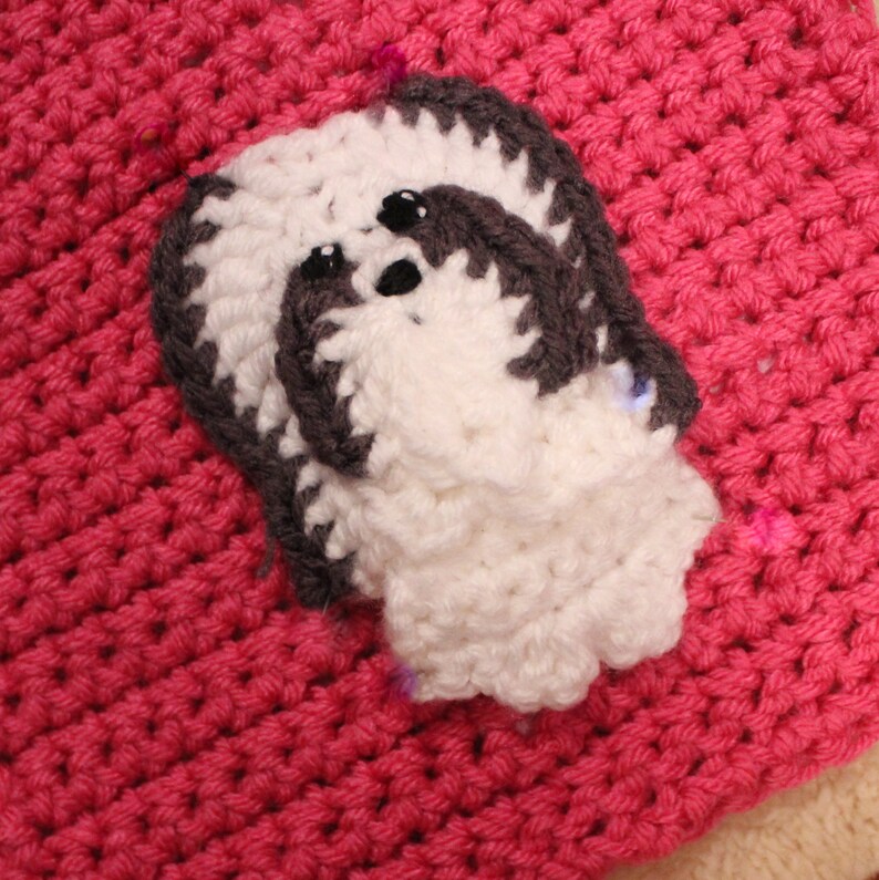 May include: A white and gray crocheted dog applique with black eyes. The applique is on a pink crocheted background.