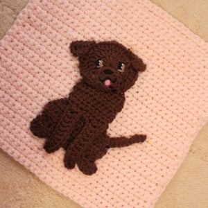 May include: A brown crocheted dog applique is pinned to a pink crocheted blanket.