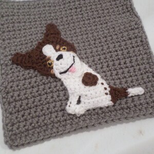 May include: A crocheted gray square with a brown and white dog applique. The dog is smiling with a pink tongue sticking out. The dog has brown spots on its white fur.