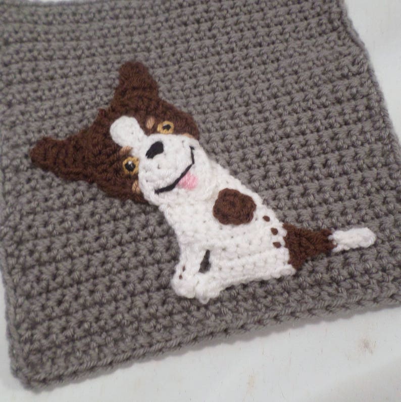 May include: A crocheted gray square with a brown and white dog applique. The dog is smiling with a pink tongue sticking out. The dog has brown spots on its white fur.