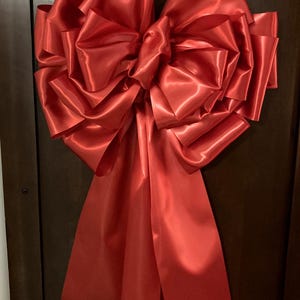 May include: A large, vibrant red satin bow, likely for decoration. The bow is attached to a dark brown door. The bow's multiple layers and long, flowing ribbons create a full, eye-catching design. The satin material has a slight sheen.