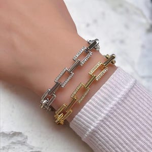 May include: Two chain bracelets with square links, one silver and one gold, both with clear cubic zirconia stones.