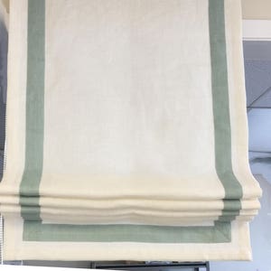 May include: A white roman shade with a green border. The shade is pulled up and folded, showing the layers of fabric.