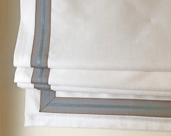 May include: White roman shade with a blue and brown trim. The shade is raised, showing the layers of fabric.