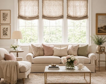 Pode incluir: Um sofá de cor creme com várias almofadas em tons de rosa, bege e verde, posicionado em frente a três janelas com cortinas romanas de linho bege. Uma mesa de centro de mármore fica à frente, com uma poltrona e mesa lateral por perto.